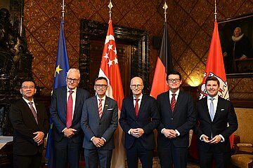 Sechs Personen in formeller Kleidung stehen in einem repräsentativen Raum vor drei Flaggen (EU, Singapur, Deutschland).