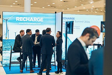 Auf einer Messe stehen mehrere Personen an einem Ausstellungsstand mit dem Schriftzug „RECHARGE“. Der Stand zeigt ein großformatiges Bild von Windkraftanlagen auf dem Meer. Die Personen tragen formelle Kleidung und unterhalten sich in kleinen Gruppen an Stehtischen. Im Vordergrund ist ein weiterer Messestand zu sehen, dessen blaues Display teilweise den Text „Global… for w…“ zeigt. Die Halle ist hell ausgeleuchtet, mit glänzendem Boden und weiteren Ständen im Hintergrund.