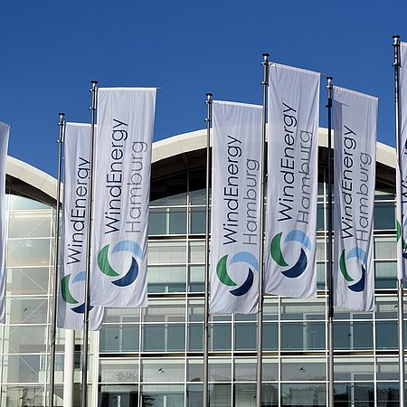 Fahnen mit dem Logo der WindEnergy Hamburg vor dem gläsernen Messegebäude bei strahlend blauem Himmel.