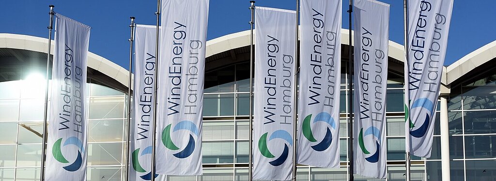 Fahnen mit dem Logo der WindEnergy Hamburg vor dem gläsernen Messegebäude bei strahlend blauem Himmel.